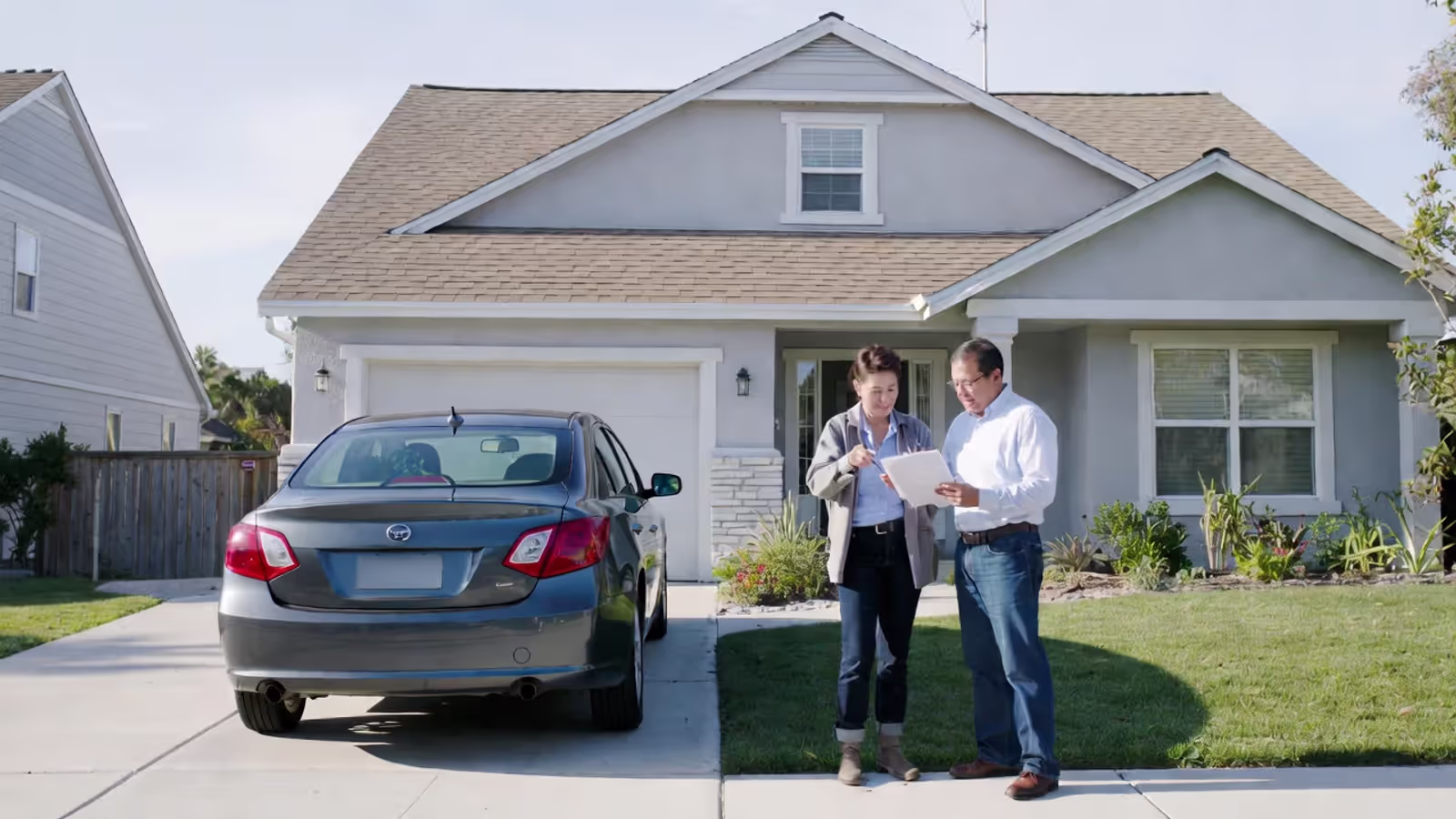 Homeowner and insurance agent reviewing an auto and home insurance policy outside a house with a car in the driveway