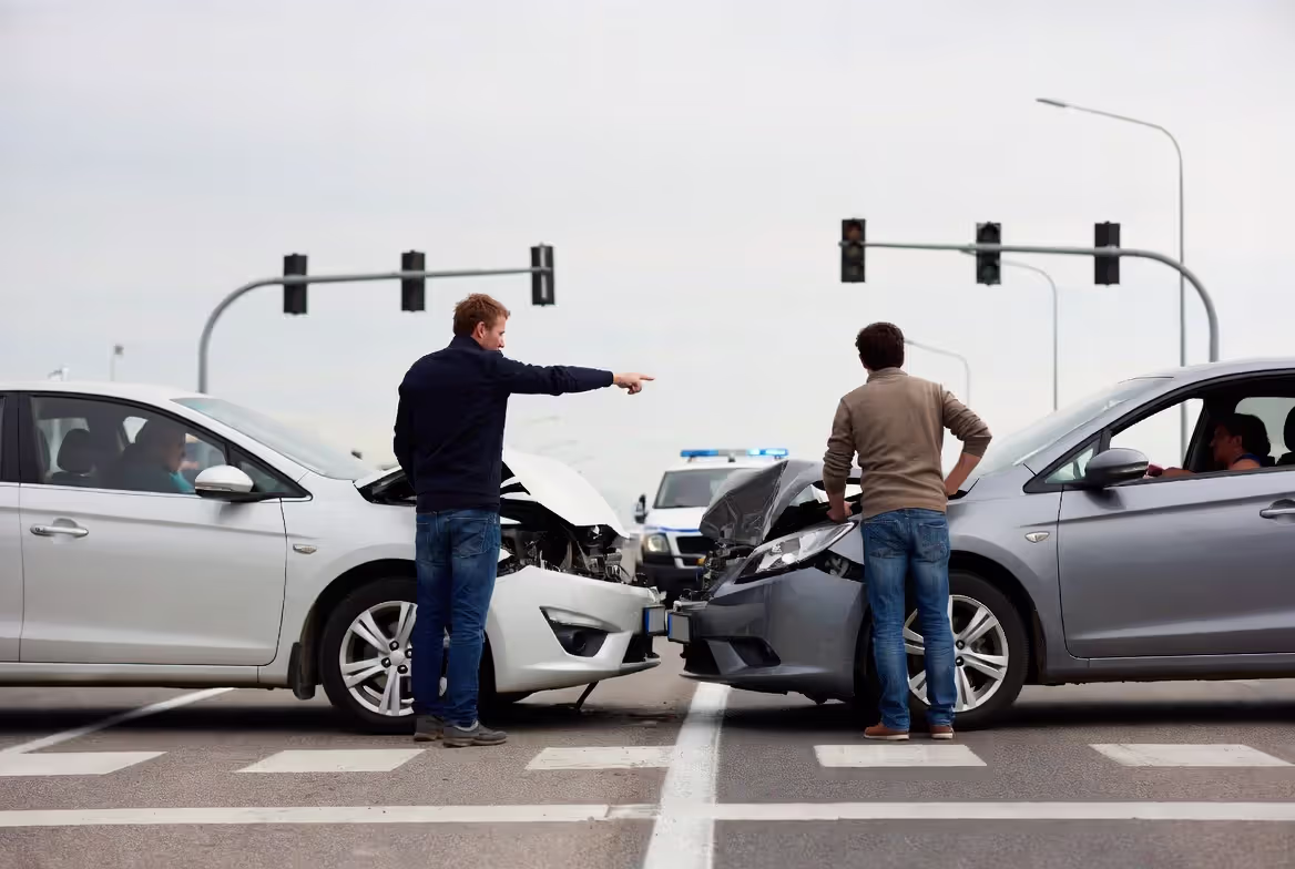 Realistic horizontal photo 16:9; two drivers standing beside damaged cars at an intersection, discussing the accident while one points toward lane markings and traffic lights; subtle police presence in background; scene communicates fault determination after a collision; overcast daylight, documentary realism, photorealistic, highly detailed; no logos; no readable text